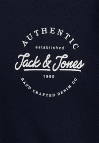 Tela de algodón de color marino con logo impreso en blanco. Presenta el texto "AUTÉNTICO" y "Jack & Jones", establecido en 1990, sobre un fondo texturizado.