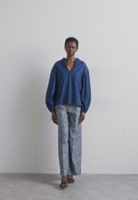 Blaut Denim-Oberteil mit V-Ausschnitt und voluminösen Ärmeln, kombiniert mit hellblauen, strukturierten, weiten Hosen. Einfache Sandalen runden den Look ab.
