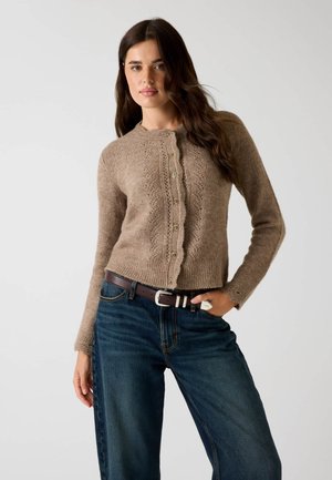 Kvinde iført en brun strikket cardigan med knapper og mørkeblå jeans med et brunt bælte, stående mod en ensfarvet lys baggrund.