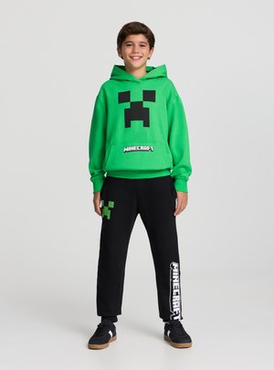 Felpa verde con grafica del volto nero del Creeper e scritta "Minecraft"; abbinata a pantaloni da ginnastica neri con dettagli verdi e bianchi.