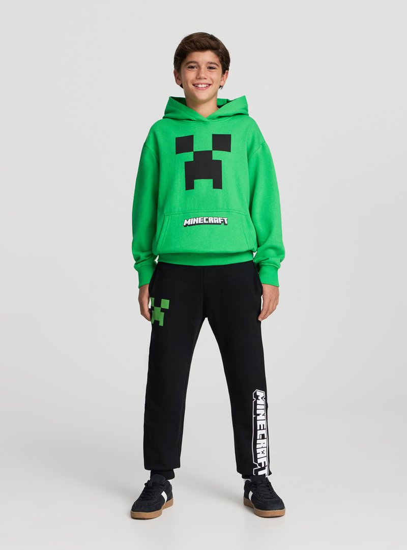Felpa verde con grafica del volto nero del Creeper e scritta "Minecraft"; abbinata a pantaloni da ginnastica neri con dettagli verdi e bianchi.