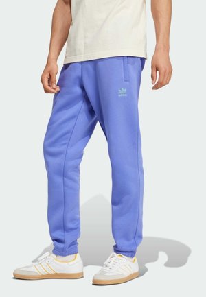 Mann trägt lavendelfarbene Adidas-Sweatpants und weiße Sneakers mit gelben Akzenten, steht vor einem schlichten hellen Hintergrund.