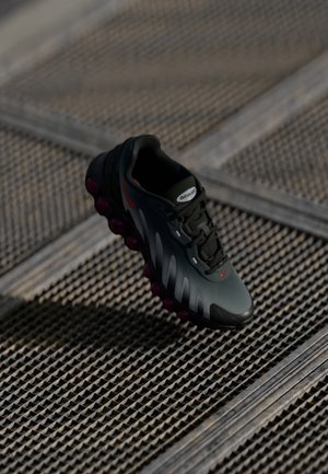 Ciemnoszary i czarny sneaker Nike Air Max z czerwonymi detalami, unoszący się nad teksturowaną metalową kratownicą w miękkim oświetleniu.