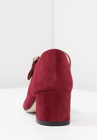 Chaussure à talon haut en daim rouge avec un talon épais et un détail de boucle sur le côté. Texture lisse, bout rond et éléments de design minimalistes.