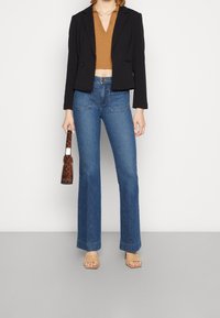 Blazer nero corto con due cerniere, top marrone a scollo a V, jeans chiari a zampa d'elefante, sandali piatti color nudo e borsa a tracolla marrone con fantasia.