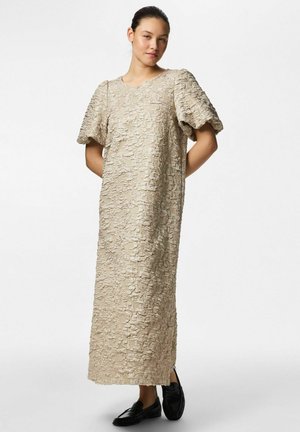 Młoda kobieta stojąca z rękami za plecami, ubrana w beżową, teksturowaną sukienkę midi z bufkami na rękawach oraz czarne mokasyny na białym tle.