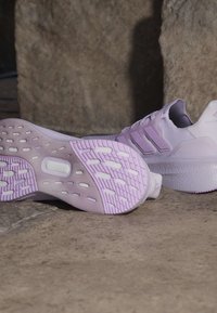 Двойка лавандулови маратонки Adidas с текстурирани подметки, поставени на каменна повърхност пред фон от груба каменна стена.