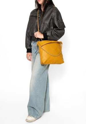 Personne portant une veste en cuir noire et un jean large bleu clair tenant un grand sac matelassé jaune moutarde avec une bandoulière chaîne.