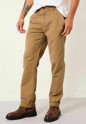Beige Baumwoll-Chinos mit geradem Bein, ausgestattet mit einem Knopfverschluss, seitlichen Taschen und aufgerollten Säumen. Getragen mit dunkelbraunen Stiefeletten.