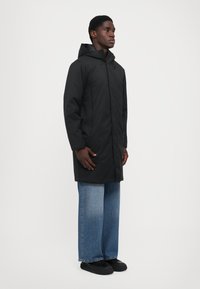 Uomo che indossa un lungo cappotto con cappuccio nero, jeans larghi blu e scarpe nere, in piedi contro uno sfondo bianco uniforme.