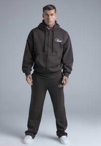 Brun zip-up hoodie och matchande mjukisbyxor i bomullstyg, med en framficka och logodetalj på bröstet och låret, kombinerat med vita sneakers.