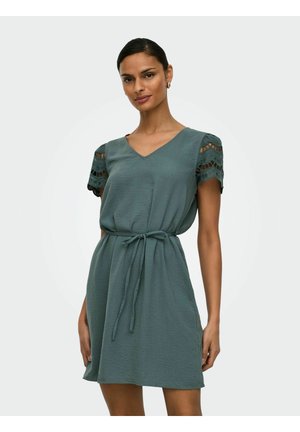 Nainen pukeutuneena merensiniseen polvipituiseen mekkoon, jossa on pitsiset lyhyet hihat ja solmittu vyötärövyö, seisoo yksinkertaista vaaleaa taustaa vasten.