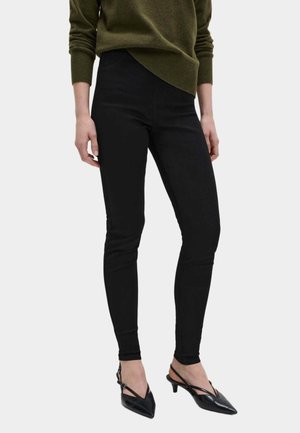 Femme portant un pantalon skinny noir, un pull vert olive et des chaussures slingback à talons aiguilles noirs, debout devant un fond uni.