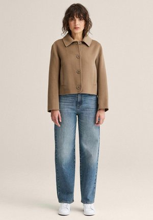 Kvinde iført en kamelfarvet jakke med knapper, blå jeans og hvide sneakers, stående mod en ensfarvet beige baggrund.