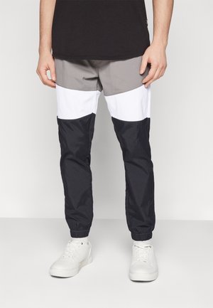 Mann trägt Jogginghose mit Farbblock in Grau, Weiß und Schwarz, kombiniert mit weißen Sneakers und einem schwarzen Shirt.