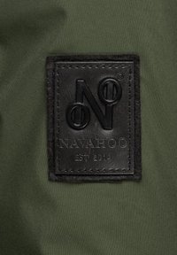 Green fabric background with a rectangular black leather patch featuring embossed "NAVAHOO" and "EST 2014" text, outlined in stitching.