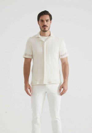 Homme aux cheveux et à la barbe foncés portant une chemise à manches courtes en tricot blanche sur un t-shirt blanc et un pantalon blanc, se tenant devant un fond uni.