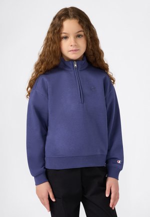 Jeune fille avec des cheveux bouclés portant un sweat-shirt à fermeture éclair violet et un pantalon noir, se tenant devant un fond blanc uni.
