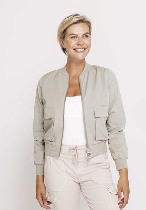 Lächelnde Frau mit kurzen blonden Haaren, die eine hellbeige Bomberjacke über einem weißen Top und passende Jogginghosen mit Kordelzug trägt, vor einem schlichten Hintergrund stehend.