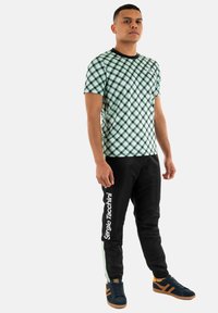 T-shirt a quadri verdi e neri con colletto tondo, abbinata a pantaloni neri con logo bianco e strisce laterali verdi. Sneakers blu.