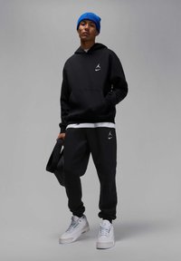 Felpa con cappuccio nera con logo bianco, pantaloni jogger neri abbinati e sneakers bianche. La persona indossa un berretto di maglia blu e tiene una borsa.