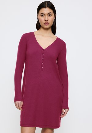 Kvinna med mörkt hår som bär en långärmad, ribbad, magenta V-ringad klänning med fem knappar framåt mot en neutral bakgrund.
