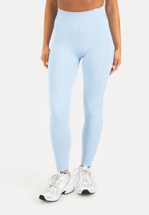 Donna che indossa leggings azzurri chiari a vita alta, scarpe da ginnastica bianche e calzini bianchi con il testo "SMILOX" su uno sfondo semplice.