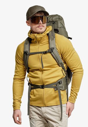 Homme portant une veste zippée jaune, un pantalon beige, une casquette verte, de grandes lunettes de soleil et portant un sac à dos de randonnée vert avec les sangles attachées.