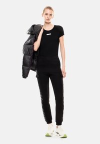 T-shirt aderente nera, pantaloni jogger neri con tasche laterali e pannelli neri lucidi, e un piumino nero appoggiato sulla spalla. Sneakers bianche.