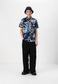 Chemise à manches courtes, couleur marine, avec un motif de feuilles tropicales colorées ; assortie à un pantalon noir large et un chapeau seau noir ; chaussures noires imposantes.
