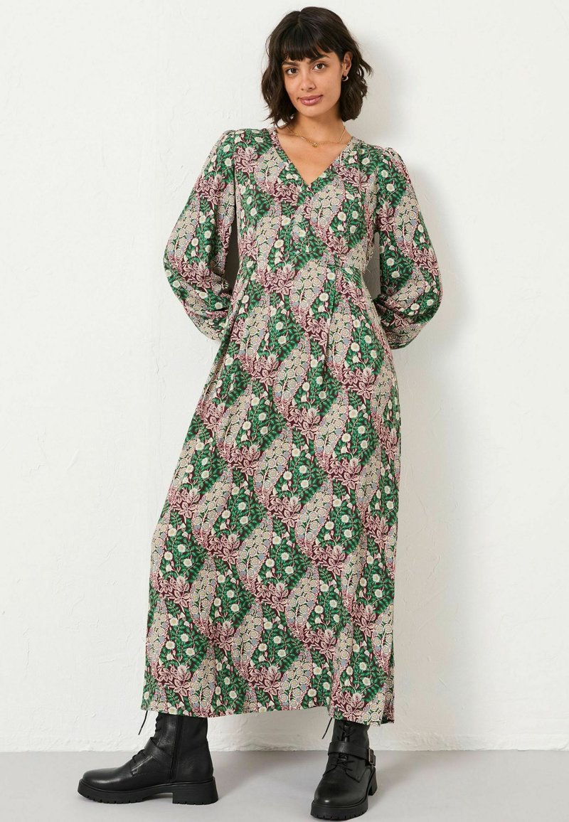 Langes, gemustertes Kleid mit tiefem V-Ausschnitt, Ballonärmeln und floralen Motiven in Grün, Pink und Beige. Kombiniert mit schwarzen Knöchelstiefeln.
