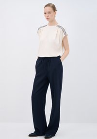 Blusa beige chiaro con perline decorative sulle spalle, abbinata a pantaloni blu scuro larghi in un tessuto testurizzato e scarpe blu scuro.