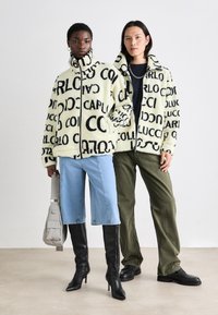 Deux personnes portent de grandes vestes en polaire ornées d'un motif de logo noir sur fond crème. Une paire de bottes à talons hauts, un short en jean et un pantalon cargo complètent les tenues.