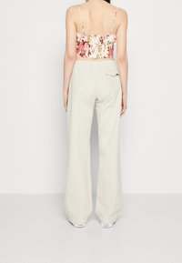 Kvinde iført en blomsteret crop top, beige vide bukser med en baglomme og hvide Converse sneakers, stående med ryggen til.