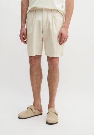 Mann trägt beige knielange Shorts, ein helles Hemd und beige Slip-On-Schuhe vor weißem Hintergrund.