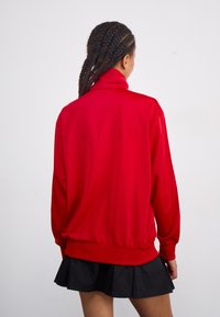 Rote Jacke mit langen Ärmeln und hohem Kragen, aus glattem Material. Hat einen lockeren Schnitt und elastische Bündchen, kombiniert mit einem schwarzen, plissierten Rock.