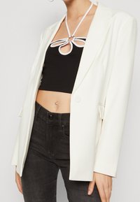 Kvinna klädd i en vit kavaj över en svart crop top med vita band, ihop med högmidjade svarta jeans och en navelpiercing.