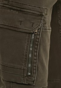 Un pantalon cargo vert olive présente une grande poche à rabat et une poche zippée en biais. Le tissu a une texture lisse avec des détails de couture visibles.