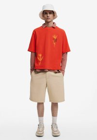 Rød bomulds T-shirt med gul blomsterbroderi, lagdelt over en langærmet pink skjorte, parret med beige shorts og en hvid bucket hat.