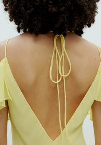 Geel chiffon jurk met een diepe V-rug, korte fladdermouwen en strikaccenten bij de halslijn, die een soepele textuur benadrukken.