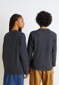 Două persoane stau cu spatele, îmbrăcate în cămăși cu manșete lungi de culoare gri închis, cu o textură uzată. Una poartă o fustă din denim, iar cealaltă are pantaloni din bumbac khaki.