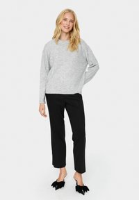 Szary sweter dzianinowy, czarne dopasowane spodnie oraz czarne buty na obcasie z ostrym noskiem i akcentem kokardki. Modelka stoi z jedną ręką na biodrze.