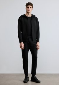 Czarna bluza z kapturem na zamek z logo, połączona z czarnymi spodniami z elastycznym pasem i czarnymi trampkami. Gładka tekstura, dopasowany krój, minimalistyczny styl.
