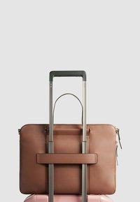 Bolsa de ordenador portátil de cuero marrón asegurada en una maleta rosa, con un asa superior y un cierre de cremallera, con una textura suave y un diseño minimalista.