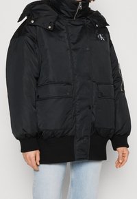 Chaqueta de plumas negra con cuello alto, cierre de cremallera y broche, dos bolsillos frontales y puños acanalados, con un parche de logo en el pecho.