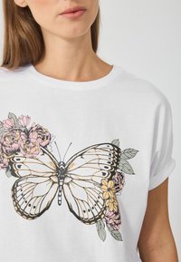 Hvid T-shirt med en stor sommerfugletegning med blomsterdetaljer i pink og gul, rund halsudskæring og rullede korte ærmer.