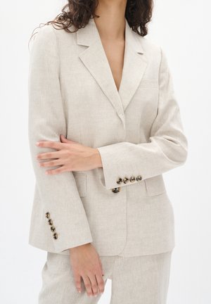 Béžový lněný blazer s klopovým límcem, jednou kapsou a šesti černými knoflíky na rukávech, s uvolněným střihem.