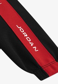 Black sportswear with a red stripe featuring "JORDAN" in white text. Smooth fabric texture, fitted design, and elastic cuffs.