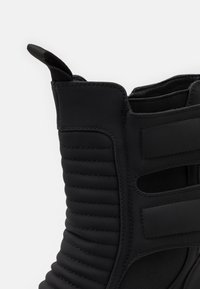 Bota negra con un acabado texturizado, acanalado vertical, panel elástico y una pestaña para facilitar el uso. Material sintético suave en general.