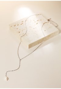 Srebrny naszyjnik łańcuszkowy z skręconym wzorem, z naturalnym akcentem w postaci perły na końcu. Prezentowany na teksturowanej płycie kamiennej.